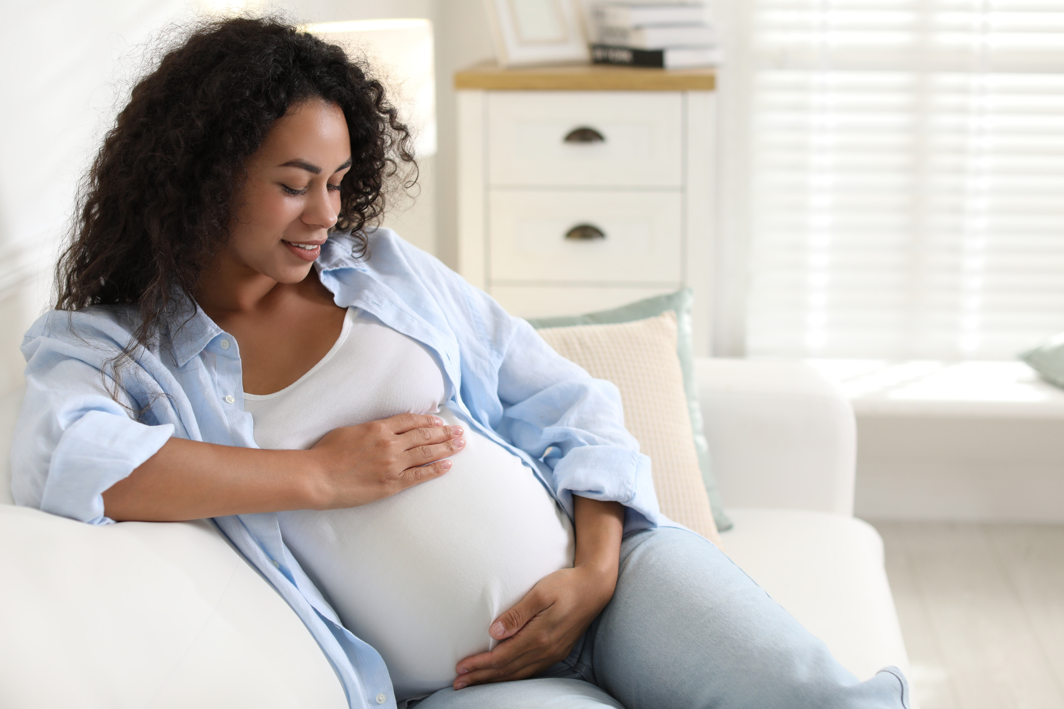 A pregnant woman sits on a couch gently cradling her belly in an image representing fetal movement monitoring, or Count the Kicks.