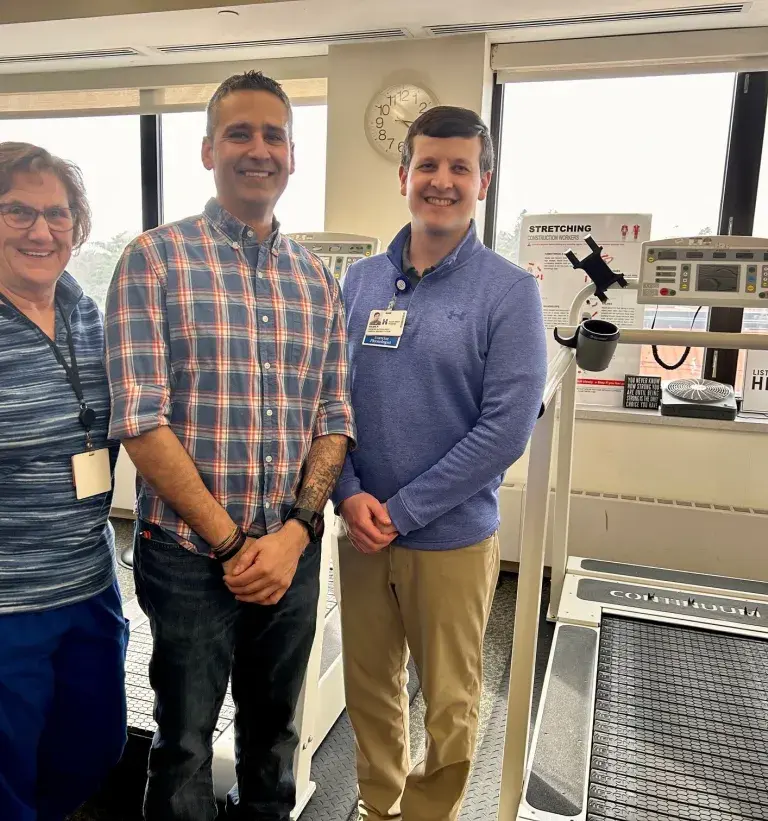 A male patient stands near a treadmill between a female nurse and male exercise physiologist
