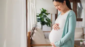 A young Asian woman smiles as she looks down at and holds her pregnant stomach.