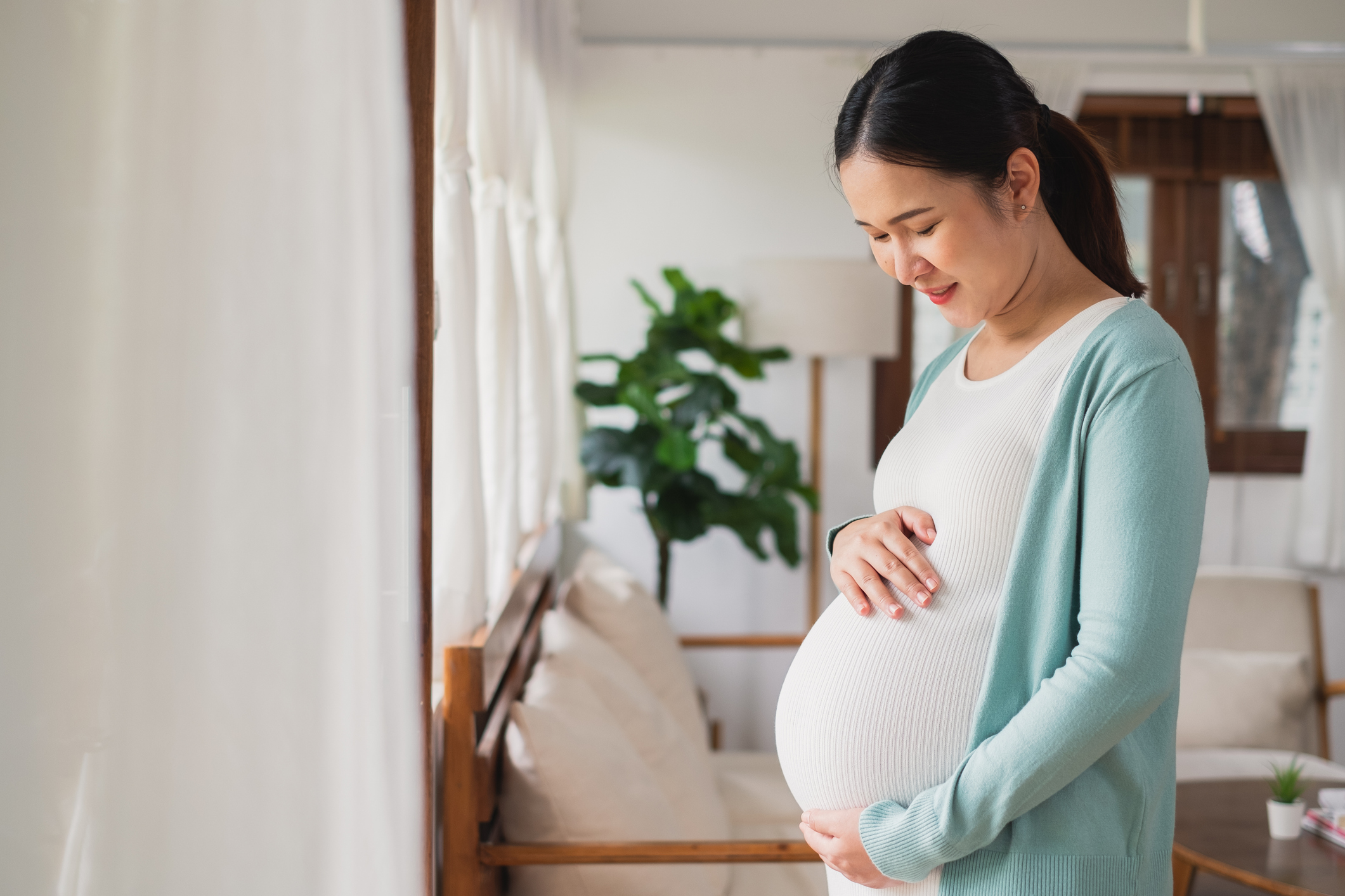 A young Asian woman smiles as she looks down at and holds her pregnant stomach.