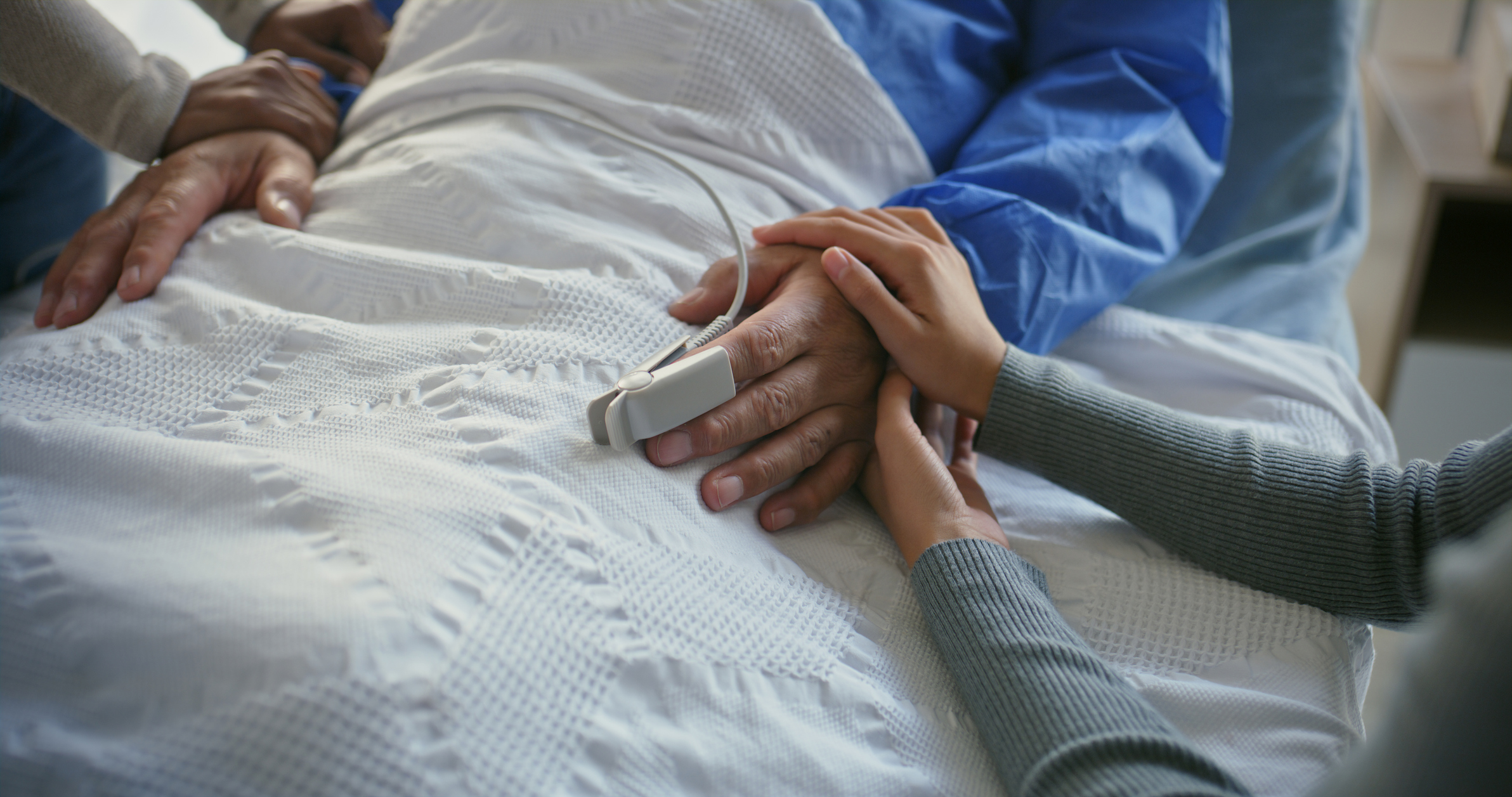 Close up view of a man's hands as he lies in a hospital bed and two people on either side of him gently place their hands on his hand and arm.