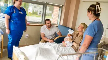 Two nurses look on as a new mom holds her infant in a hospital bed at South Shore Hospital with her husband next to her.