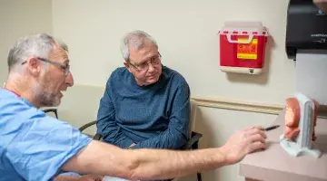 Sergio Fefer, MD points to an anatomical model as he speaks to an older patient seated in a chair.